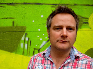 A man with short brown hair wearing a red and white plaid shirt against a bright green graffiti background.