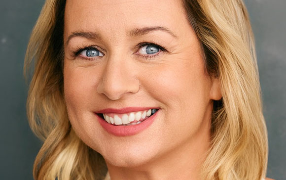 A smiling woman with blonde hair and blue eyes looking toward the camera.