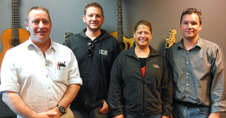 Four adults standing side by side in a room with acoustic and electric guitars hanging on the wall.