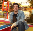 A man wearing a denim jacket and black shirt sits on a colorful boat near a canal with other boats.