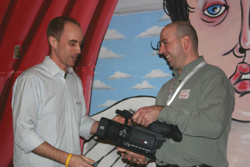Two men stand in front of a colorful mural, one holding a professional video camera.