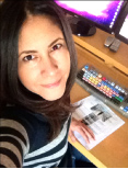 A woman with dark hair smiles while sitting at a desk with a computer, keyboard, and papers.