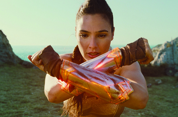 A woman crosses her glowing, metallic gold gauntlets in front of her chest in a defensive pose.