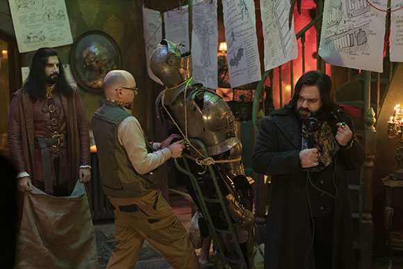 Three men stand in a room filled with sketches and a suit of armor.