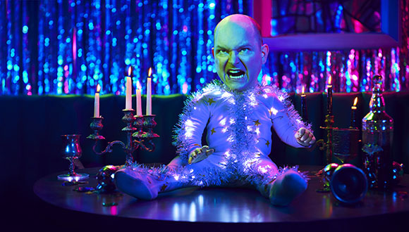 A small, bald, expressive figure in a white sequined bodysuit sits at a table with lit candles.