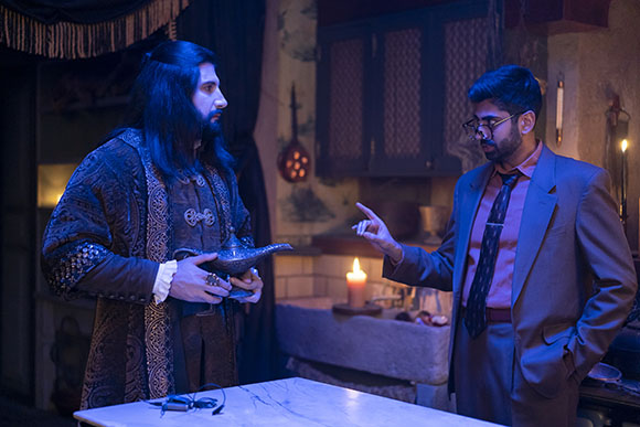 A man in a dark patterned robe holds a genie lamp while gesturing to a man in a suit and glasses.