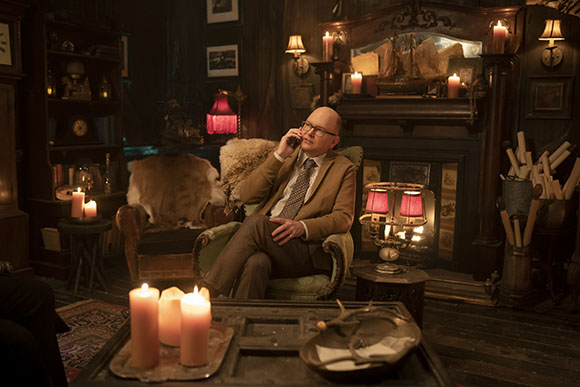 A man in a brown suit sits in an armchair, talking on a mobile phone in a room filled with candles.