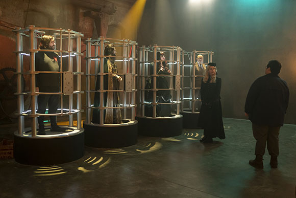 Four people stand inside glowing metal cylindrical cages while two others watch in a dark, moody room.