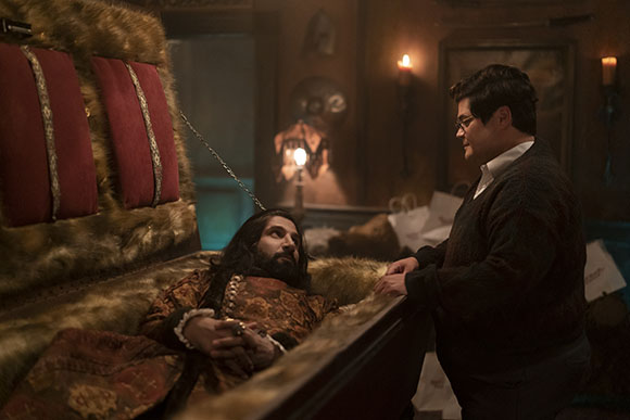 A man in a dark sweater stands next to a man with long dark hair lying in a fur-lined wooden coffin.
