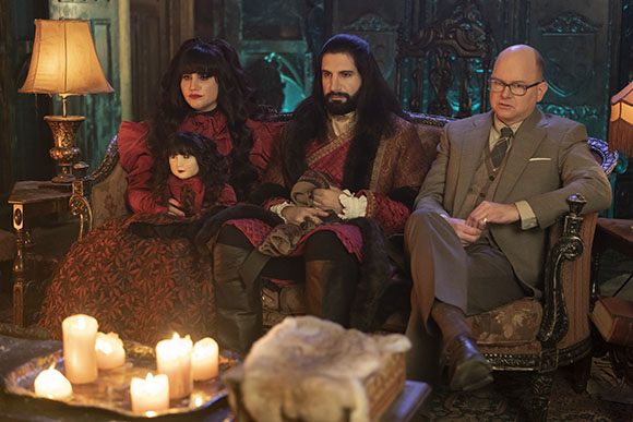 Three people in historical costumes sit on an ornate sofa near lit candles.
