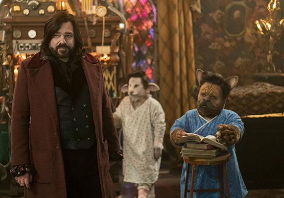 A man in a burgundy coat stands in a workshop alongside two anthropomorphic creatures.
