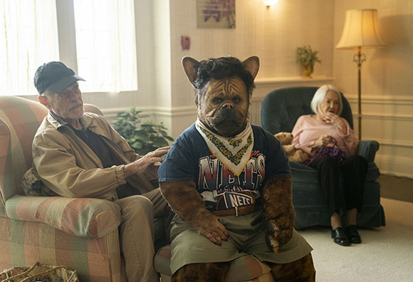 A person in a brown furry creature mask and costume sits between an elderly man and woman in a living room.