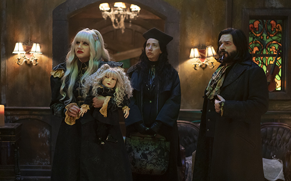 Three people in dark, gothic clothing stand in a dimly lit room with stained glass and candlelight.