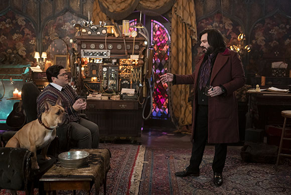 A man in a long maroon coat gestures toward a seated man and a tan dog in a room filled with vintage equipment.