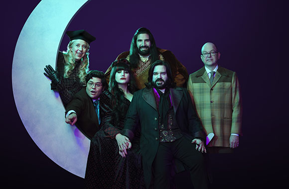 Six people in theatrical costumes pose around a large white crescent moon against a dark purple background.