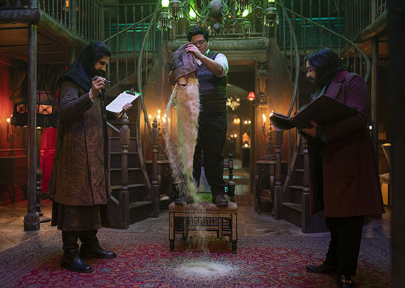 Three men in a dark, decorated room watch as a substance pours from a bag held by a man in the center.