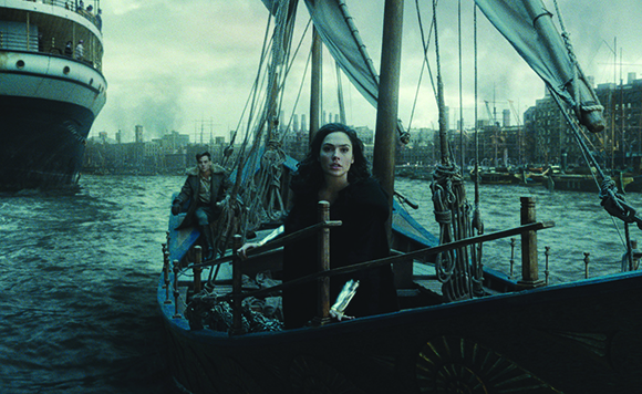 A woman in black stands on a small wooden sailboat in a harbor near a large ocean liner.