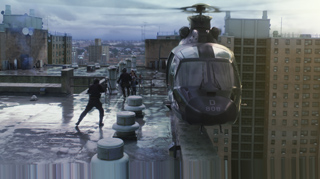 A dark helicopter hovers beside a city rooftop where people are standing near the edge.