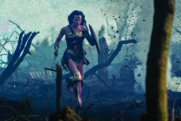 A woman in superhero armor runs through a dark, smoky forest landscape with debris in the air.