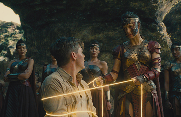 A woman in golden armor holds a glowing lasso around a man in a tan shirt in a rocky setting.