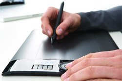 A person's hands using a black stylus on a digital graphics tablet over a white surface.