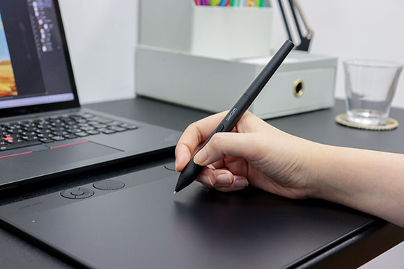 A person's hand holds a black Wacom stylus against a digital drawing tablet near a laptop.