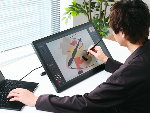 A person uses a stylus on a large digital drawing tablet connected to a laptop on a white desk.