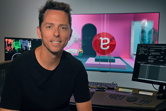 A man in a black t-shirt sits before a large screen displaying colorful graphics and a professional editing suite.