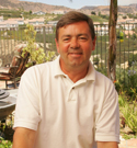 A smiling man wearing a white polo shirt sits outdoors with a hilly residential landscape in the background.