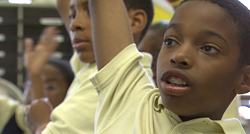 Two young boys in light yellow shirts raising their hands in a classroom setting.