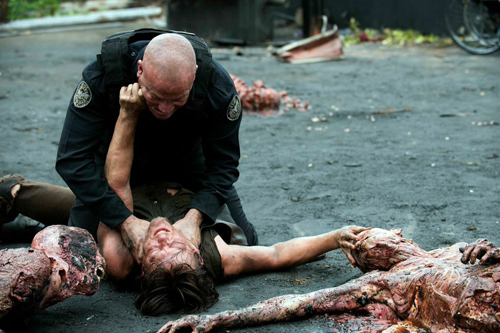 A police officer pins a person to the ground surrounded by several bloody corpses on an asphalt surface.