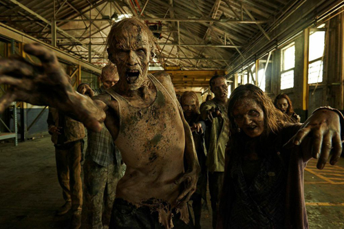A group of decaying zombies with outstretched hands reaching toward the viewer in a dark warehouse.
