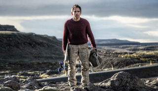 A man in a maroon sweater and tan pants stands in a vast, rocky landscape under a cloudy sky.