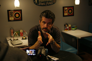 A man with dark hair and a beard gestures with both hands while sitting at a table.