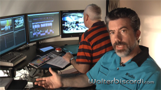 A man in a blue shirt gestures in the foreground while a technician works at multiple monitors in the background.