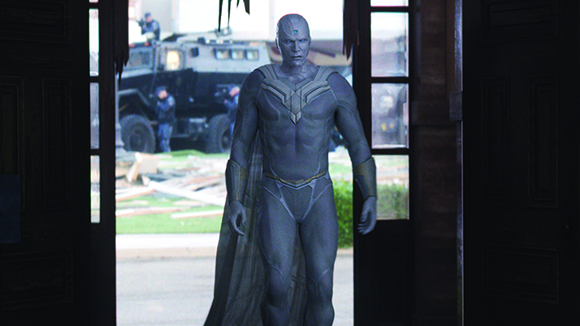 A muscular man in a blue bodysuit and cape stands in a doorway with an armored vehicle in the background.