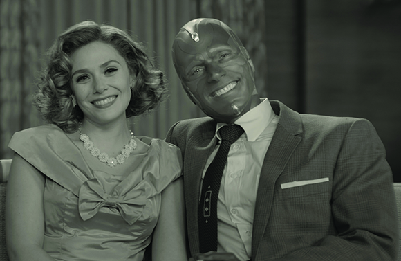 A smiling woman in a vintage dress and a man wearing a superhero mask and suit pose for a photo.