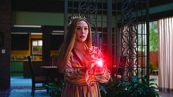 A woman in a striped dress holds her hands out with a bright red magical glow between her palms.
