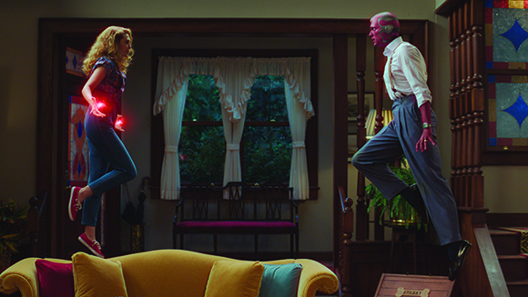 A woman with blonde hair and a red-skinned man float in mid-air facing each other in a living room.
