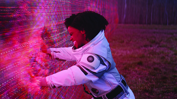 A person in a white futuristic suit reaches toward a glowing pink digital wall in a dark field.