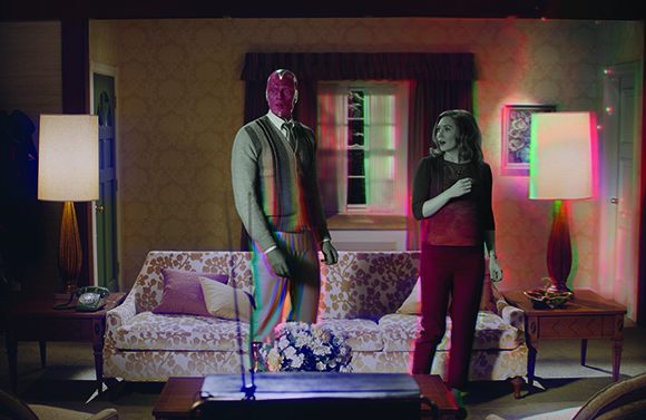 A man with red skin and a woman stand in a living room with a colorful glitch effect.