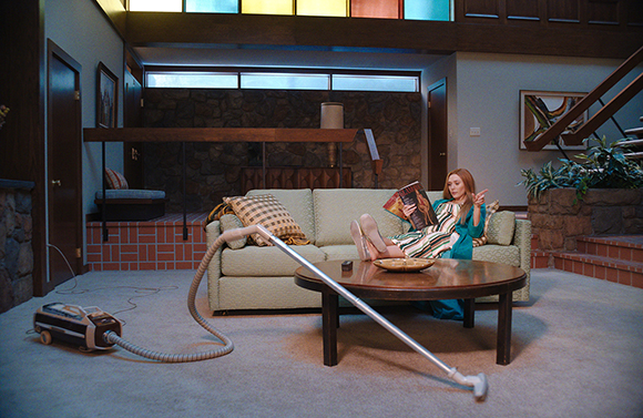 A woman in a blue dress sits on a light green sofa reading a magazine in a mid-century modern living room.