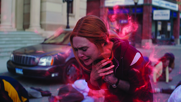 A distressed woman with red hair clutches her chest as pink smoke swirls around her in a city street.