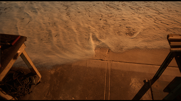 Foamy ocean waves wash over a concrete edge near wooden structures under warm lighting.