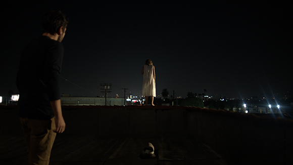 A man in a dark shirt watches a woman in a white dress standing on a distant rooftop at night.