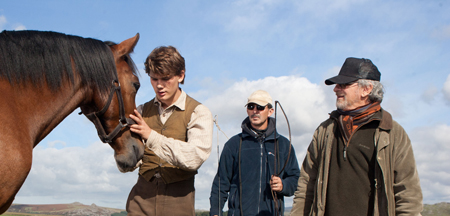 A young man in a vest pets a brown horse while two men stand nearby under a cloudy sky.