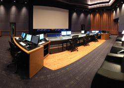 A large media control room featuring a long console with multiple computer monitors and a large white screen.