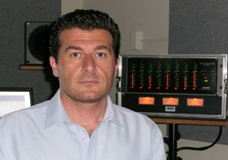 A man in a light blue shirt stands in front of audio equipment in a studio.
