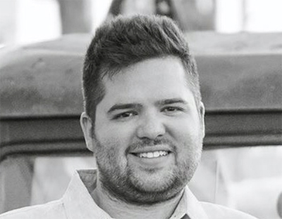 A smiling man with short dark hair and a beard, wearing a collared shirt, in a black and white portrait.