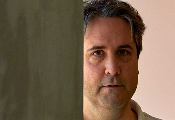 A close-up portrait of a man looking forward, partially obscured by a grey wall on the left.